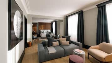 Una habitación de hotel moderna con un sofá gris, un sillón beige y una pequeña mesa redonda. Hay un ventanal con cortinas oscuras, un cuadro en la pared y suelo de madera.
