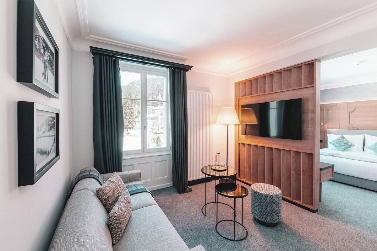 A modern hotel room with a sofa, a TV mounted on a wooden panel, a lamp, and a small round table.