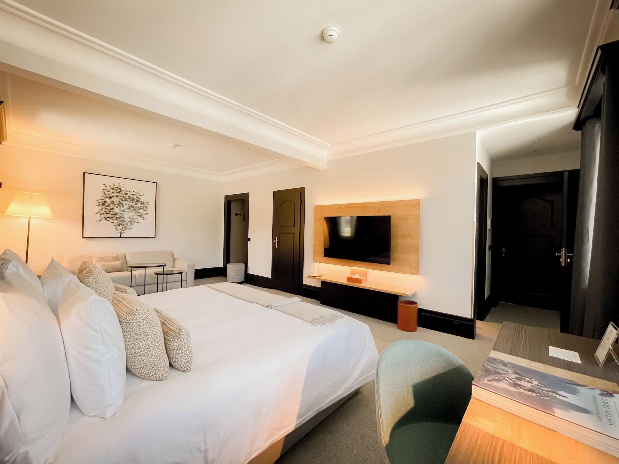 A hotel room with a large bed, a desk, a chair, a TV mounted on the wall, and a framed picture on the wall.