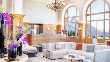 Un salon spacieux avec un grand canapé, une table basse et un lustre. De grandes fenêtres offrent une vue sur les montagnes, et on aperçoit un coin cuisine au fond de la pièce.