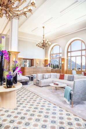 A spacious living room with a large sofa, a coffee table, and a chandelier. There are large windows with a mountain view, and a kitchen area is visible in the background.