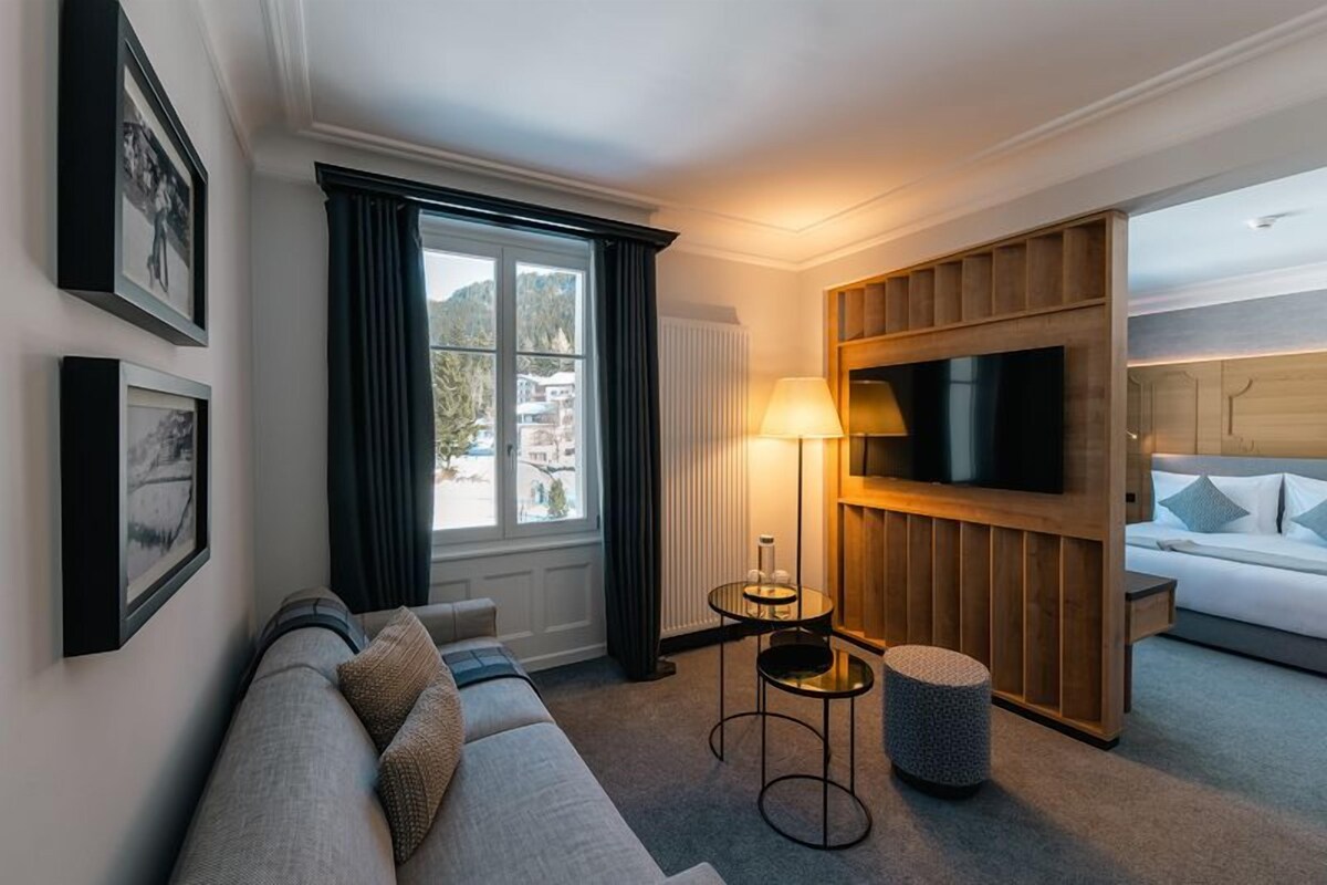 A modern hotel room with a sofa, a TV mounted on a wooden panel, a lamp, and a view of the outside.