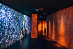 Un couloir avec un mur texturé sur le côté gauche et un mur lisse sur le côté droit, qui mène à une pièce éclairée par une source de lumière chaleureuse.
