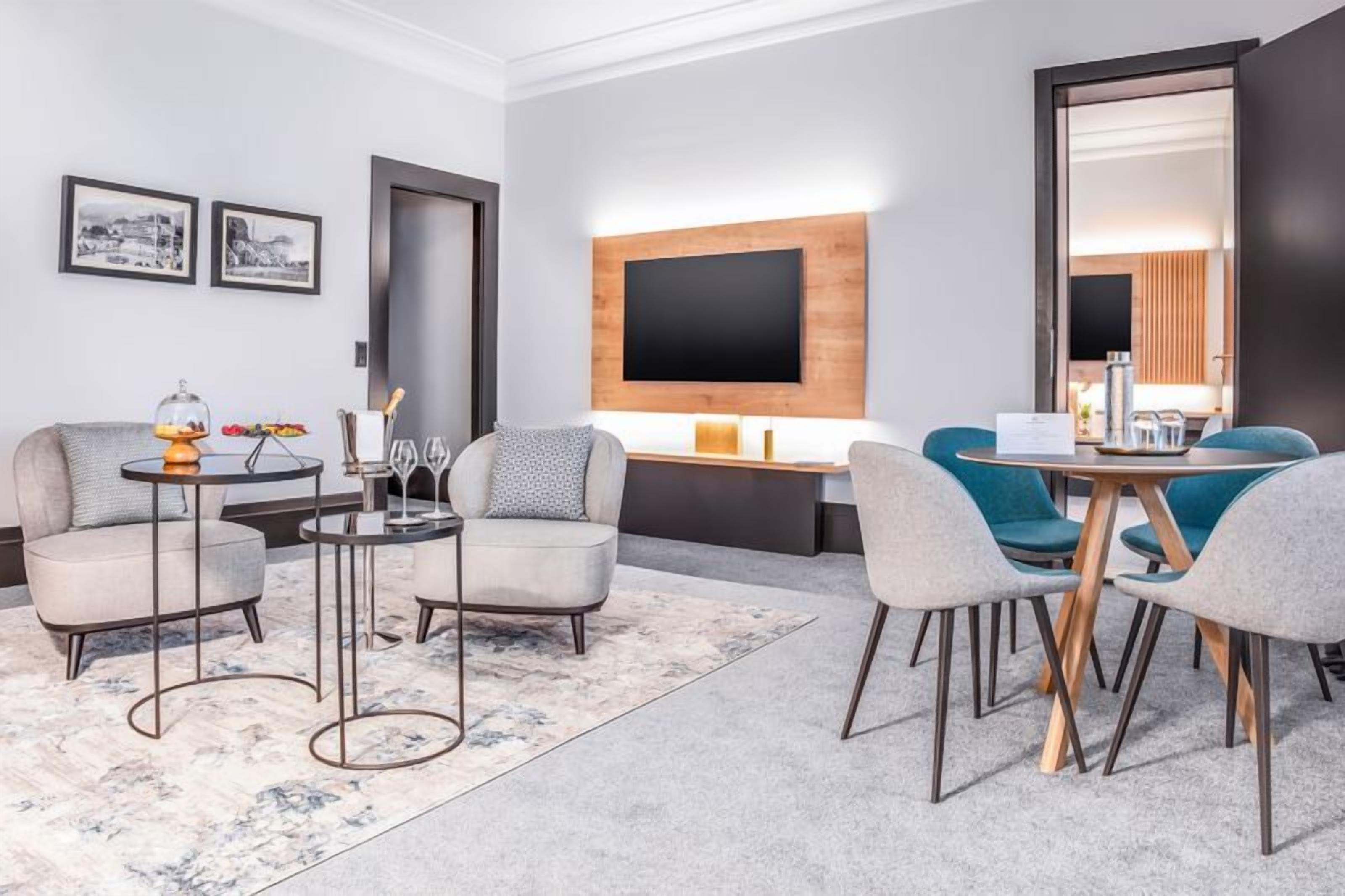 A modern hotel room with a sofa, two armchairs, a coffee table, a TV mounted on the wall, and a large mirror.