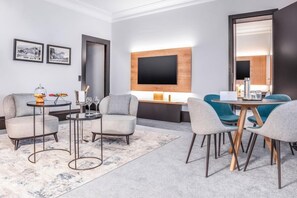 A modern hotel room with a sofa, two armchairs, a coffee table, a TV mounted on the wall, and a large mirror.