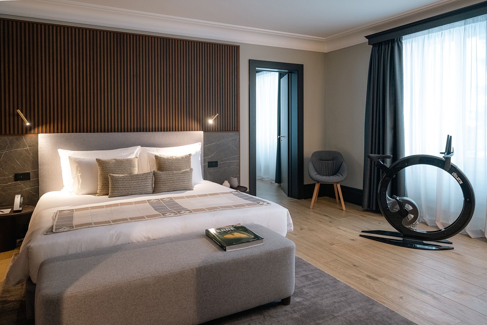 Ein modernes Hotelzimmer mit einem großen Bett, einem Stehtrainer, einem grauen Sessel und Blick ins Freie durch das Fenster.