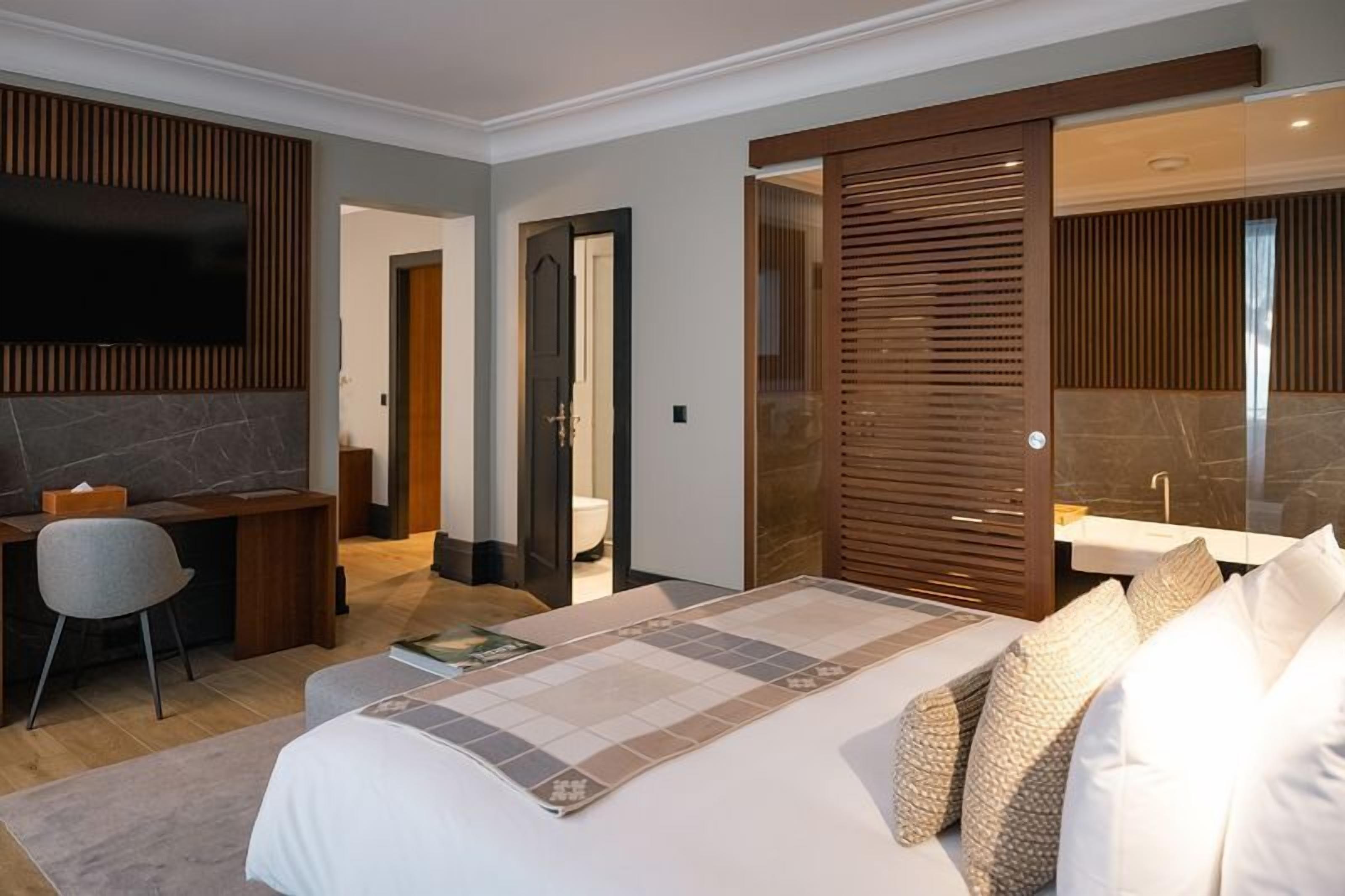 A modern hotel room with a large bed, a desk, a chair, a TV, and a bathroom with a mirror and sink.