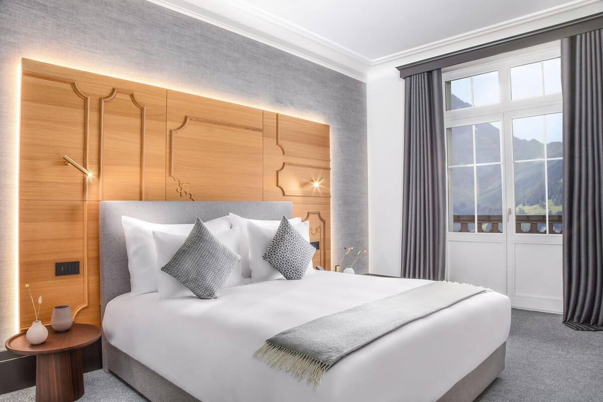 A modern hotel room with a large bed, a wooden headboard with integrated lighting, a nightstand, and a view of mountains through a large window with curtains.