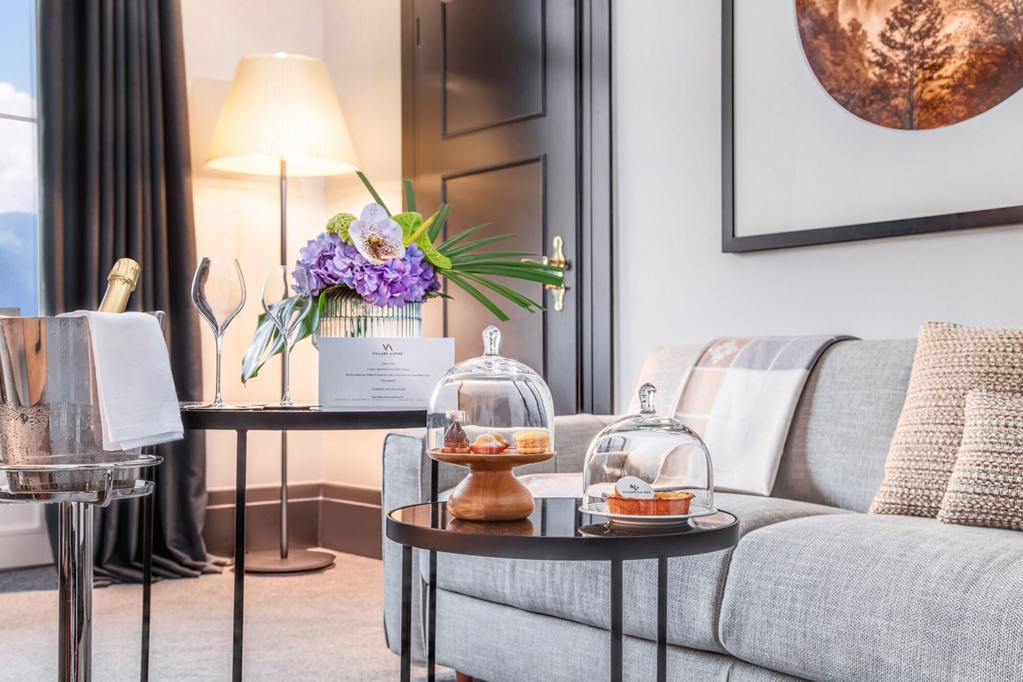 A modern hotel room with a sofa, a small table with a cake and a glass dome, a lamp, and a framed picture on the wall.