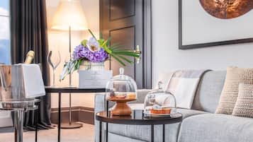 A modern hotel room with a sofa, a small table with a cake and a glass dome, a lamp, and a framed picture on the wall.