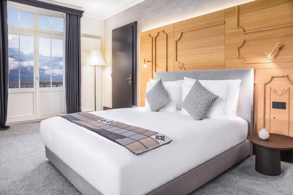 A hotel room with a large bed, a wooden headboard, a nightstand, and a view of mountains through the window.