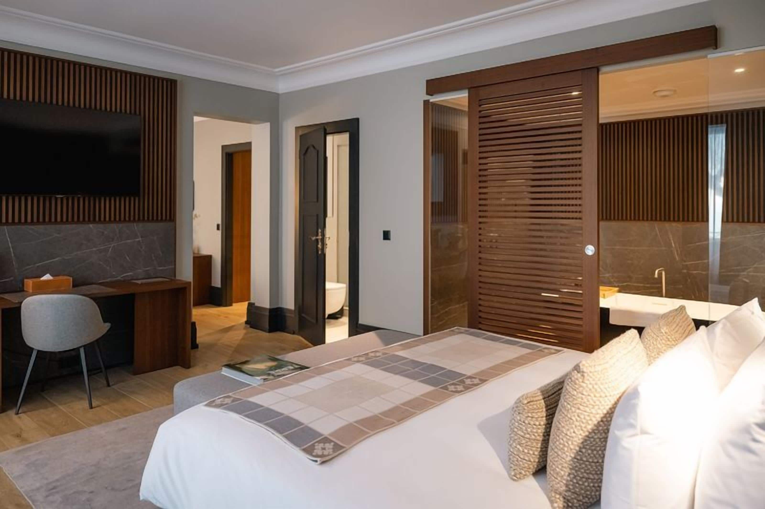 Ein modernes Hotelzimmer mit einem großen Bett, einem Schreibtisch, einem Sessel, einem Fernseher und einem Badezimmer mit Spiegel und Waschbecken.