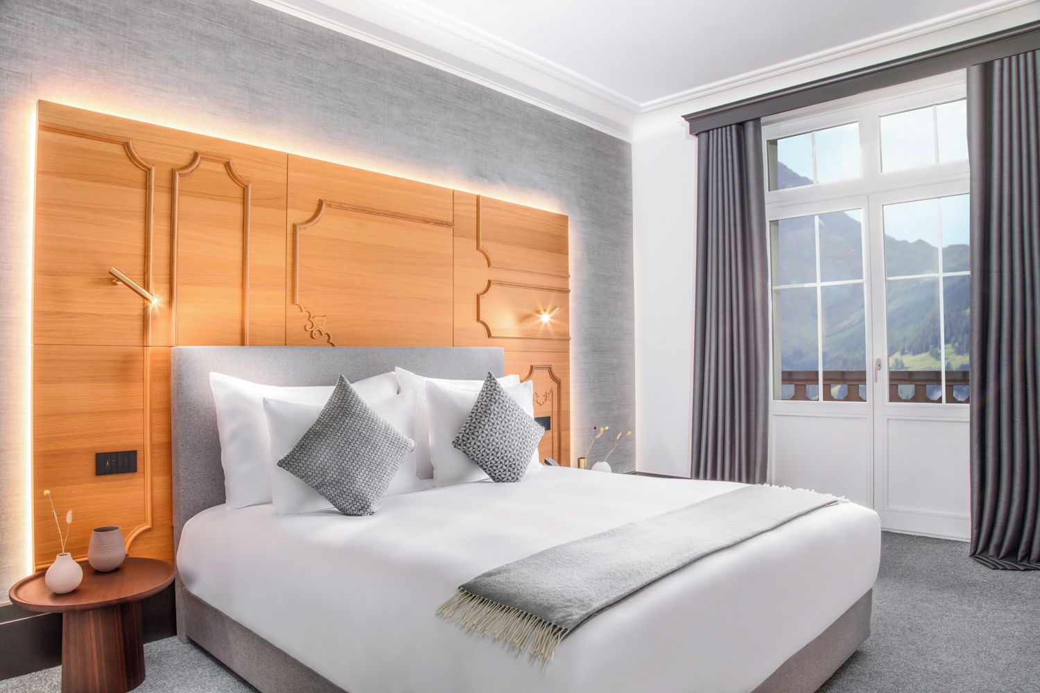 A modern hotel room with a large bed, a wooden headboard, and a view of mountains through the window.