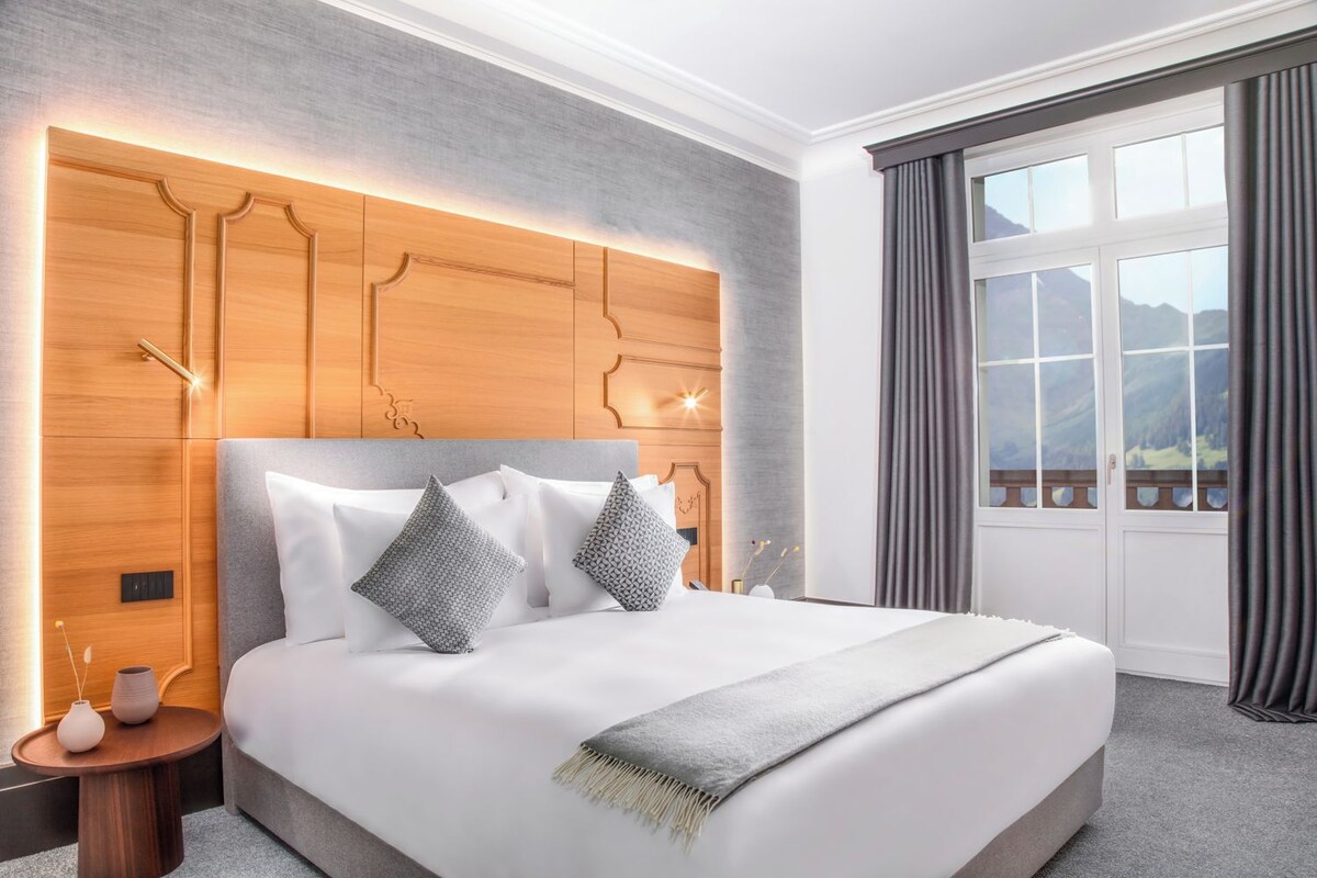 A modern hotel room with a large bed, a wooden headboard, and a view of mountains through the window.