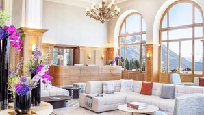 Ein geräumiges Wohnzimmer mit einer großen Couch, einem Couchtisch und einer Chandelier. Große Fenster bieten Blick auf die Berge, und im Hintergrund ist eine Küchenzeile erkennbar.