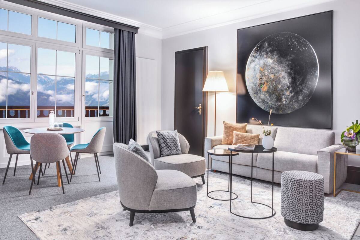 A modern living room with a sofa, armchairs, a coffee table, and a dining area with chairs and a table. There is a large window with a view of mountains and a large artwork on the wall.