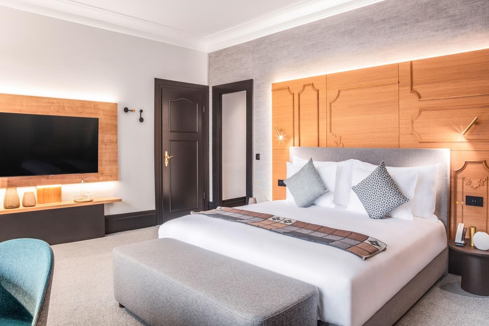 A modern hotel room with a large bed, a TV mounted on the wall, a wooden headboard, and a bedside table with a lamp.