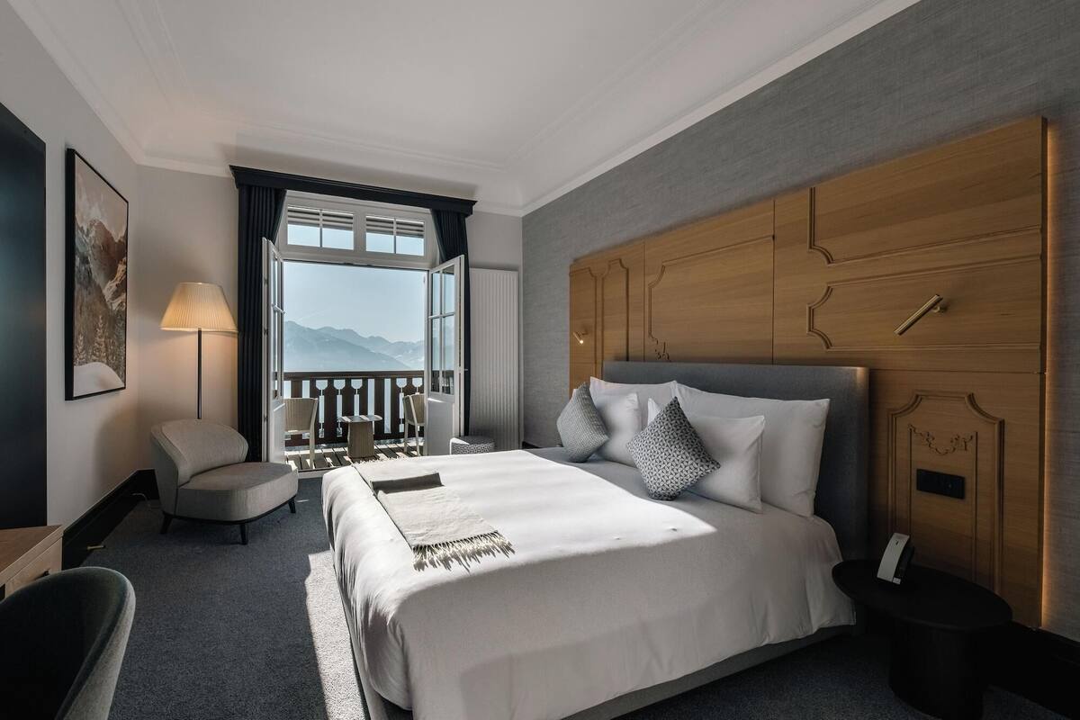 A hotel room with a large bed, a wooden headboard, a balcony with a view of mountains, a chair, and a lamp.