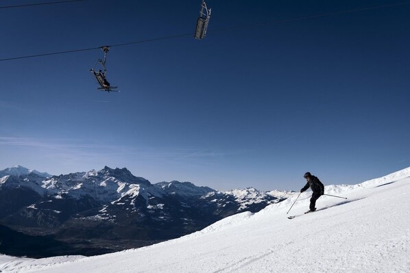 Une personne fait du ski sur une pente enneigée, avec une chaîne de montagnes en arrière-plan et un télésiège visible dans le ciel.