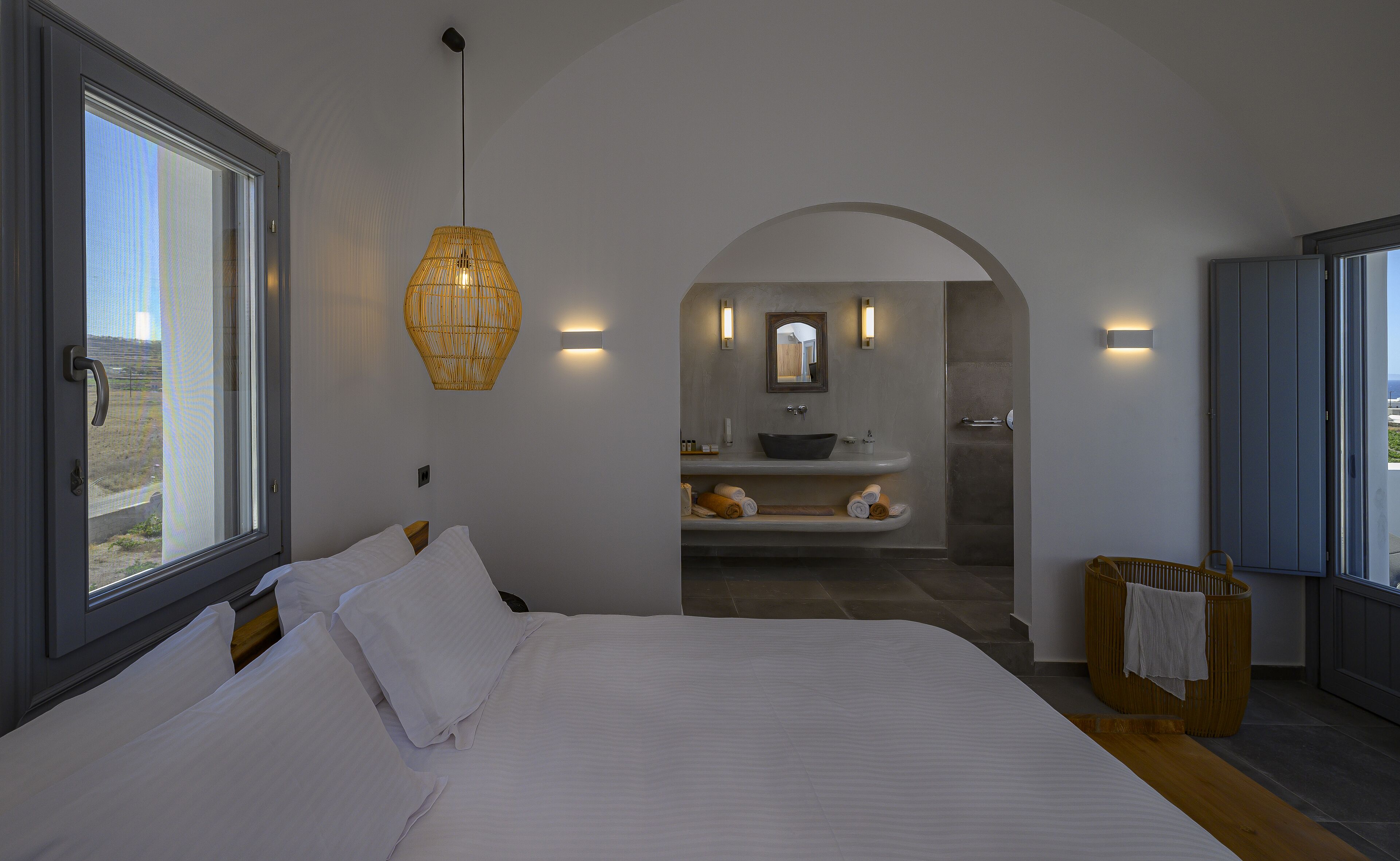 Un dormitorio con una cama grande, una ventana y vistas a un baño con lavamanos y espejo.