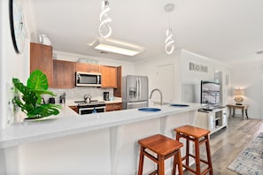 Una cocina moderna con armarios blancos, una isla central con taburetes, microondas, refrigerador y cafetera.