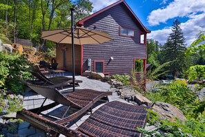 A rustic cabin with a patio area featuring wicker lounge chairs and a table, surrounded by rocks and greenery. - Rockledge Lookout (Bolton Landing)
