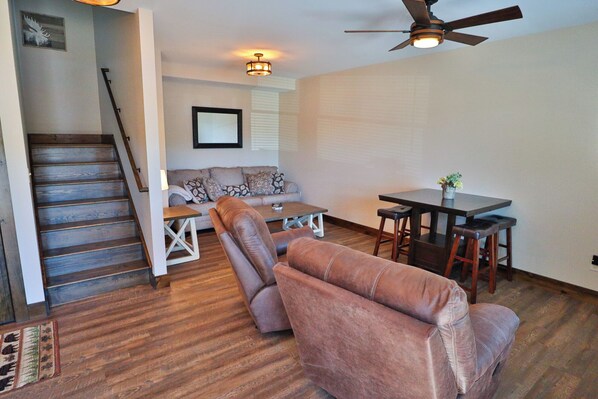 A living room with a sofa, two armchairs, a dining table with stools, a ceiling fan, and a staircase. - Rockledge Lookout (Bolton Landing)