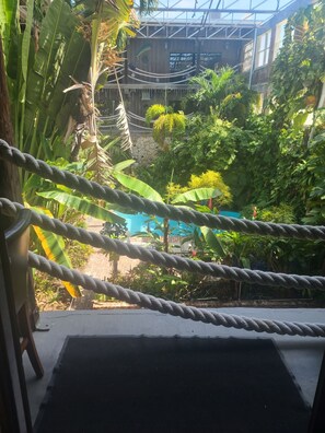Un pont de corde enjambant un bassin où prospèrent de luxuriantes plantes tropicales, avec un bâtiment dont la façade est ornée d’un symbole de poisson, visible au fond.