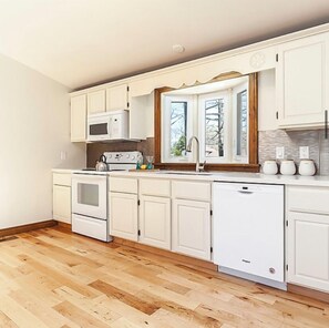 Una cocina moderna con armarios blancos, horno empotrado, microondas y lavavajillas, una ventana con vistas a árboles, y piso de madera.