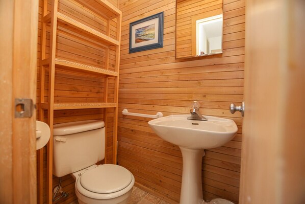 Une petite salle de bain aux murs lambrissés, équipée de toilettes, d’un lavabo et d’étagères.