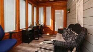 Interior acogedor de una cabaña con techo de madera, ventilador de techo, zona de comedor con mesa y sillas, sofá de mimbre y un ventanal con persianas.