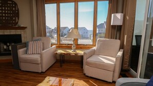 Una sala de estar con dos sillones color beige, una mesita de madera con una lámpara y vistas a casas a través de ventanas amplias.