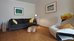 A modern living room with a dark grey sofa, a white bed with yellow pillows, a wooden coffee table, and a bookshelf.
