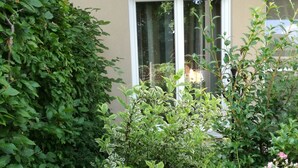 A garden with dense foliage and blooming pink and white flowers in front of a building with a window. - Luxurious apartment (2 rooms; 70 sqm; terrace; garden; air conditioning...) (Buggingen)