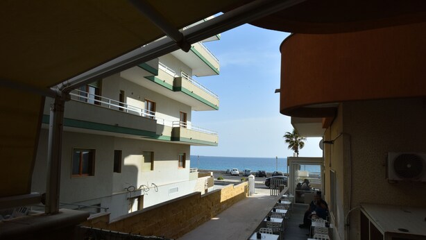 Blick auf ein Gebäude mit mehreren Balkonen, das auf einen Strand überblickt. Es gibt Außenbereiche zum Sitzen mit Tischen und Stühlen.