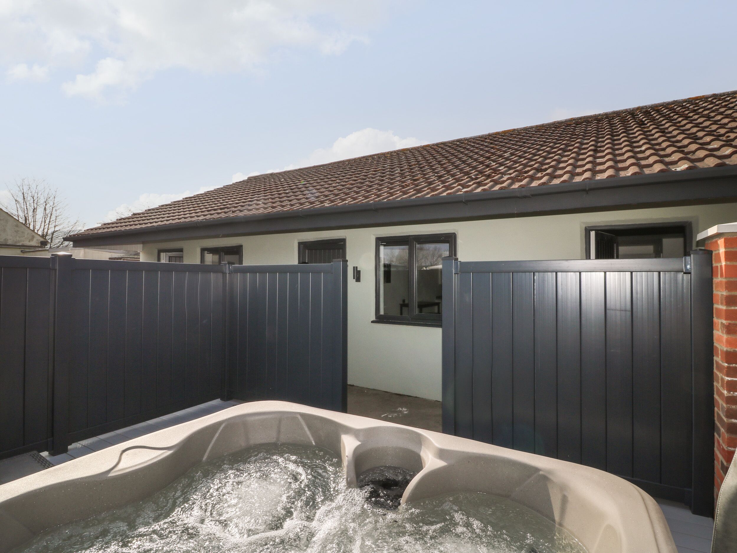 A backyard with a hot tub, a fenced area, and a house with a tiled roof.
