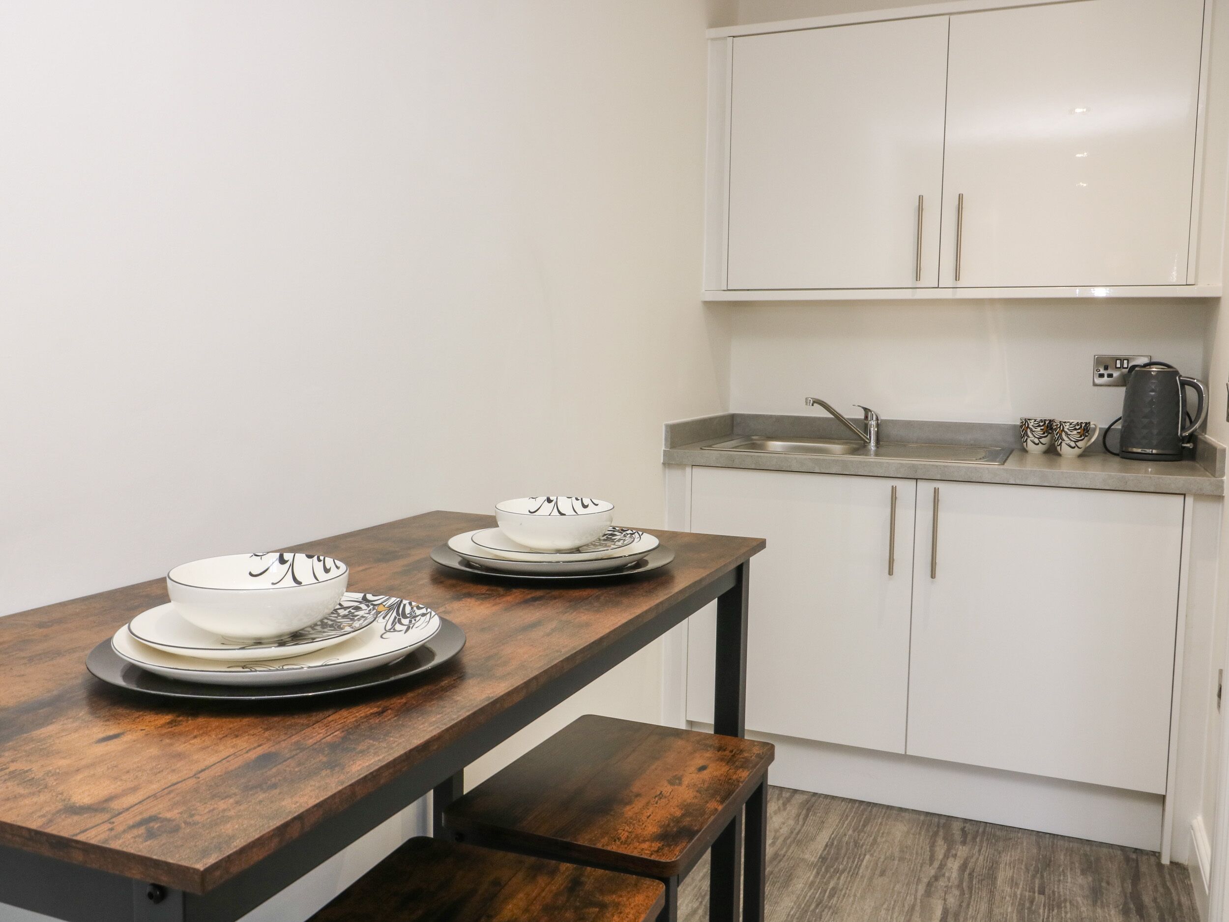 A compact kitchen with white cabinets, a wooden table with plates, and a kettle.