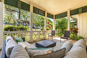 A covered outdoor seating area with wicker furniture, a coffee table, and a view of lush greenery and trees. - Home Away from Home, Beach Villa! (Kapolei)