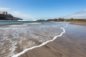 A beach with waves lapping onto the shore, a rocky outcrop in the background, and buildings on the far side.