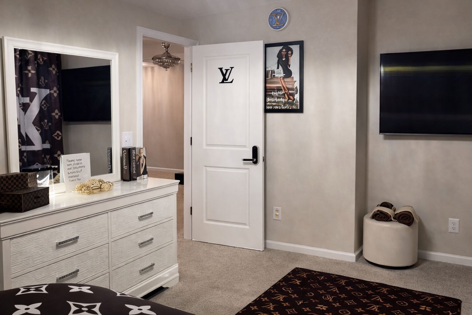 A hotel room with a white wardrobe, a Louis Vuitton sign on the door, a framed poster on the wall, and a TV mounted on the wall.