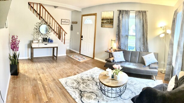 A living room with a grey sofa, a coffee table with flowers, a wooden staircase, and a door with a mirror above it. - Cheerful residential house with all yours needs. (St. Louis)