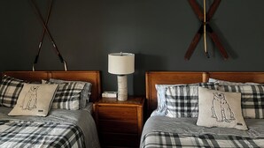 A hotel room with two beds, each with a checkered bedspread and pillows featuring a polar bear design. There is a nightstand between the beds with a lamp and a small box. The walls are dark green, and there are two wooden poles mounted on the wall.