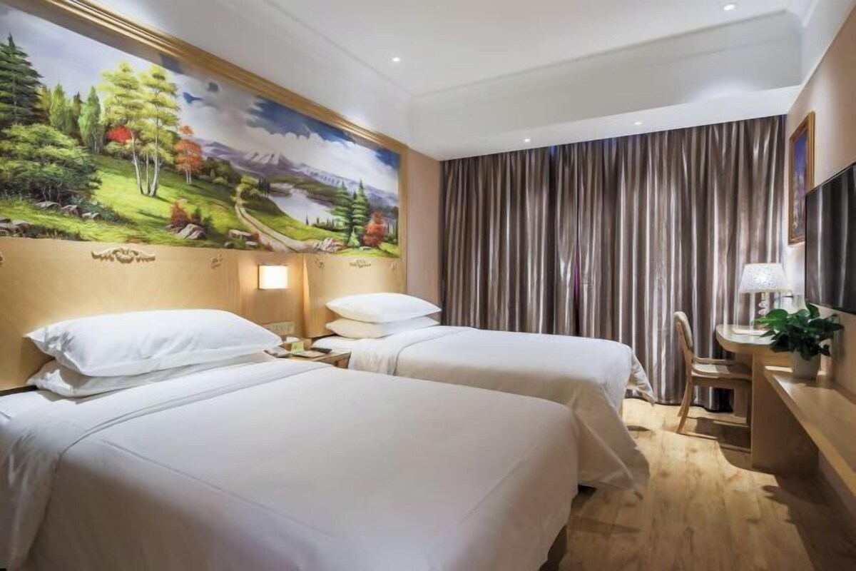 A hotel room with two beds, a large mural on the wall, a desk with a lamp, and a curtain.