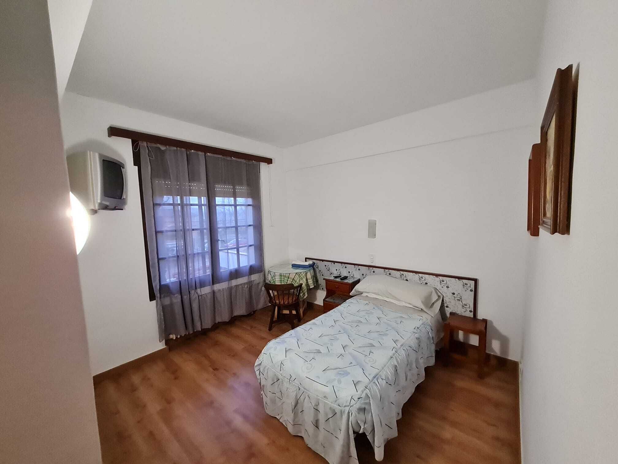 A bedroom with a bed, a small table, a chair, a TV, and a window with curtains.