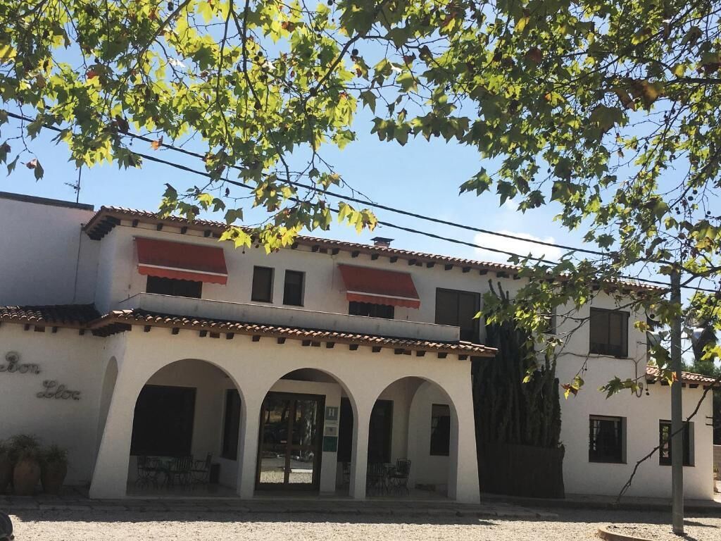 A two-story building with a white facade, red awnings, and a tiled roof. There are three arches at the ground level, and a tree with green leaves is in the foreground.
