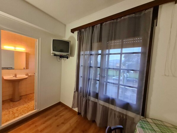 Una habitación con piso de madera, ventana con cortinas, televisor montado en la pared y un lavamanos con espejo.