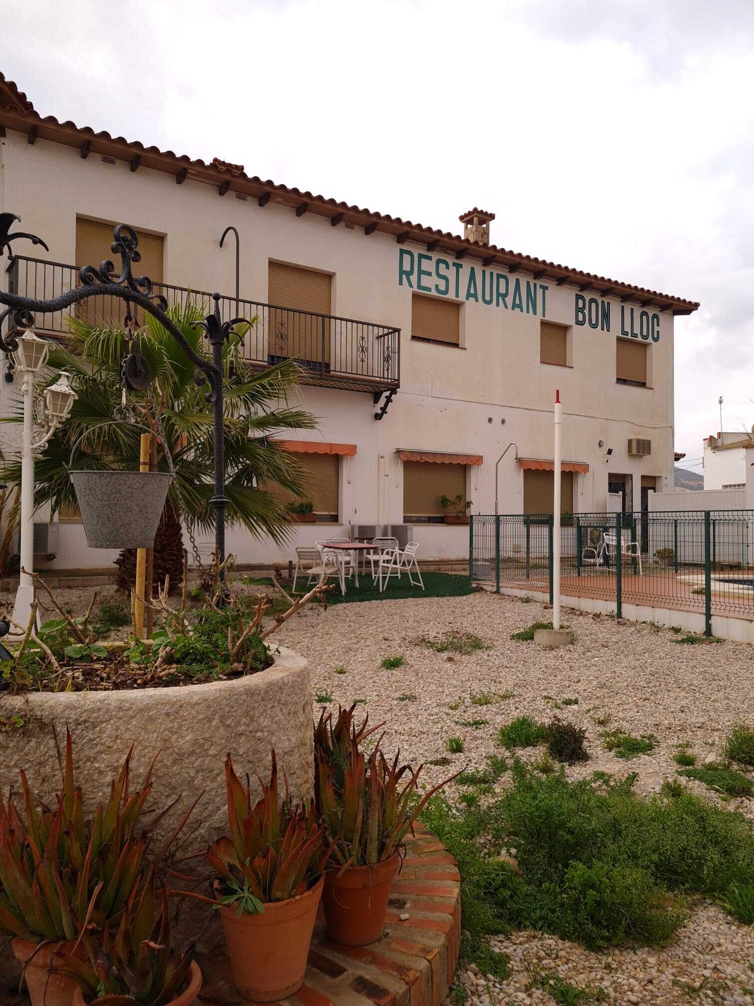 Ein Gebäude mit der Aufschrift „RESTAURANT BON LLOC“ und Außenbereich mit Sitzgelegenheiten und Blumentöpfen.