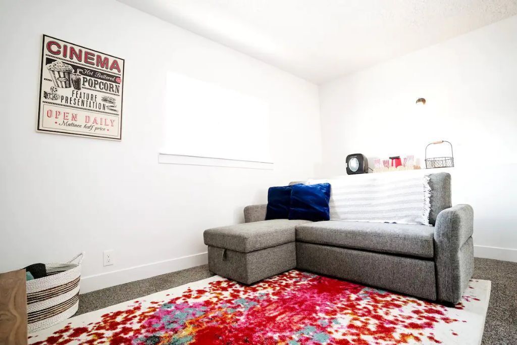 Ein Eckzimmer mit einem grauen Ecksofa, einem bunten Teppich und einem Filmplakat an der Wand.