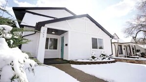 Een wit huis met een zwart dak, een voordeur en een kleine tuin met planten onder een laag sneeuw.