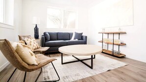 Een moderne woonkamer met een bank, een rond bijzettafeltje en een stoel. Er is een raam, een lamp en een plank met een bord.